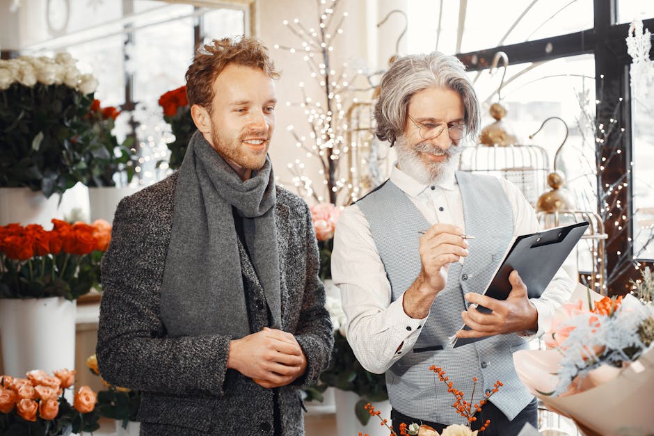 A florist and a male customer in a bright flower shop with large windows, displaying an arrangement of fresh pink, white, and orange flowers wrapped in light paper. The florist, an older man with gray