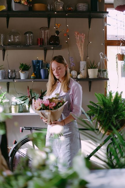 A young woman with light brown hair wearing a white shirt and apron, holding a bouquet of fresh flowers wrapped in light brown paper, featuring pink roses, white lilies, and green foliage, in a well-l