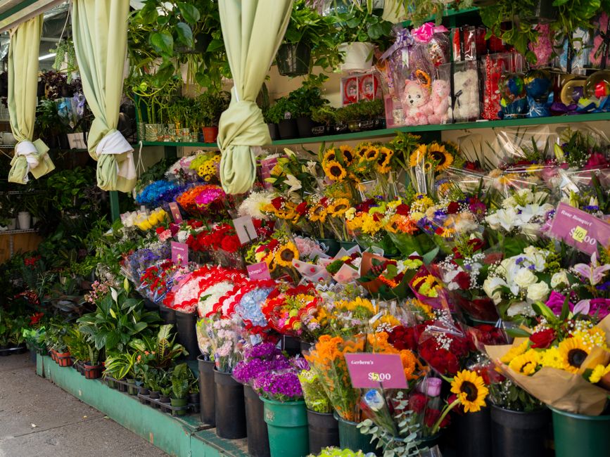 A vibrant flower display outside a florist shop features a variety of fresh floral arrangements, including bright yellow sunflowers, red roses, purple and pink carnations, white lilies, and mixed seas