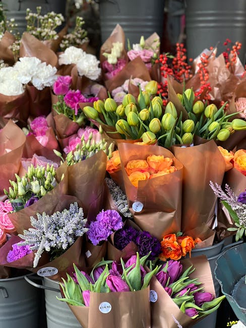 A vibrant display of freshly cut flowers arranged in brown wrapping paper, featuring a variety of blooms including white hydrangeas, pink and purple carnations, orange roses, green tulips still in bud