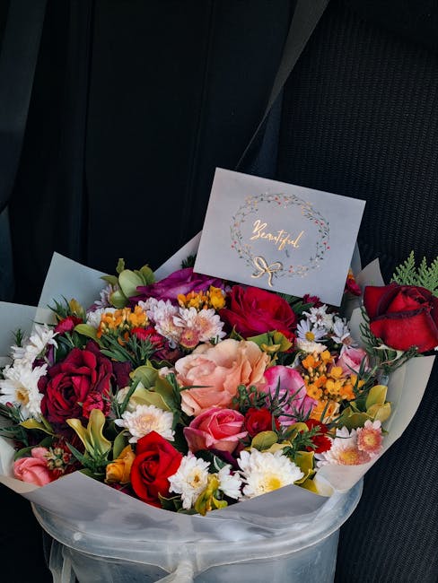 A vibrant floral bouquet arranged with a variety of fresh flowers including red roses, pink roses, peach carnations, white daisies, purple hypericum, and small orange clusters, all nestled in white wr
