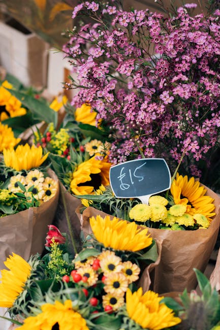 A vibrant display of fresh floral arrangements at a flower market, featuring bright yellow sunflowers, small daisy-like blossoms with cream petals and dark centers, and red buds, all wrapped in brown