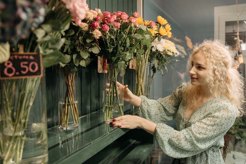A woman with curly blonde hair and a floral patterned dress arranging a bouquet of fresh flowers in tall glass vases on a green shelf at a flower shop. The bouquet features pink roses, yellow chrysant